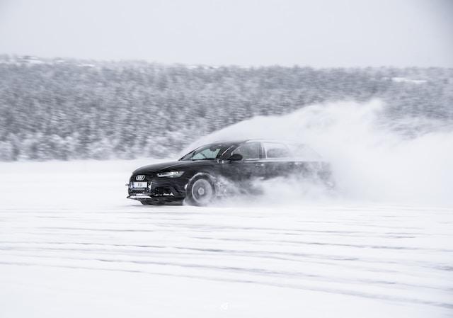 Ein schwarzer Audi, der mit hoher Geschwindigkeit über Glatteis fährt und dabei Schnee aufwirbelt, mit einem dichten Wald im Hintergrund.